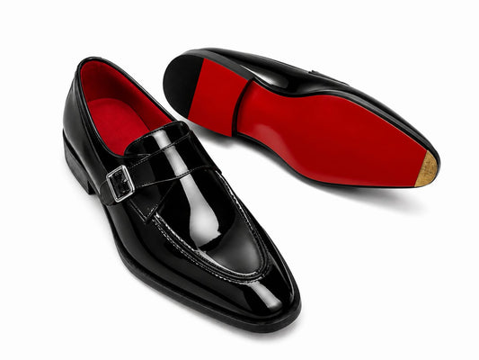 A pair of glossy black patent leather loafers with a buckle strap, shown on a white background. One shoe is positioned upright to show the black leather upper and vibrant red interior lining, while the other is turned upside down to display the contrasting bright red outsole.